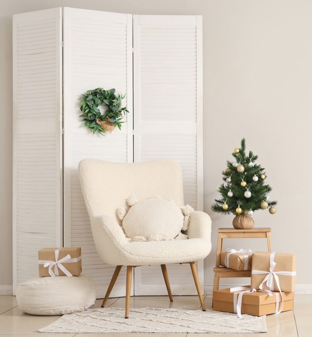 Interior of room with armchair, folding screen, small Christmas tree and gift boxes on stepladder near white wall